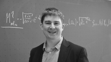 Simon Moser steht vor einer Tafel mit physikalischen Gleichungen, ausgezeichnet für gute Lehre.